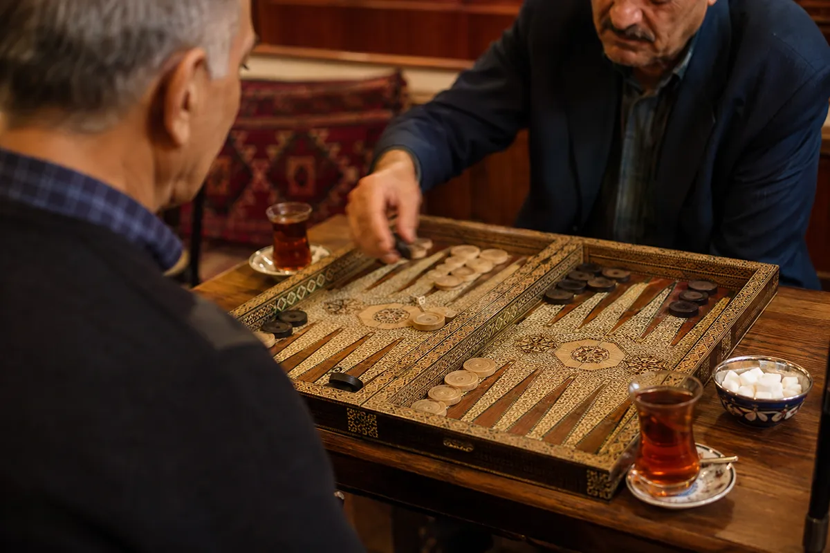 Two men playing takhteh nard at a table with glasses of Persian tea beside the board